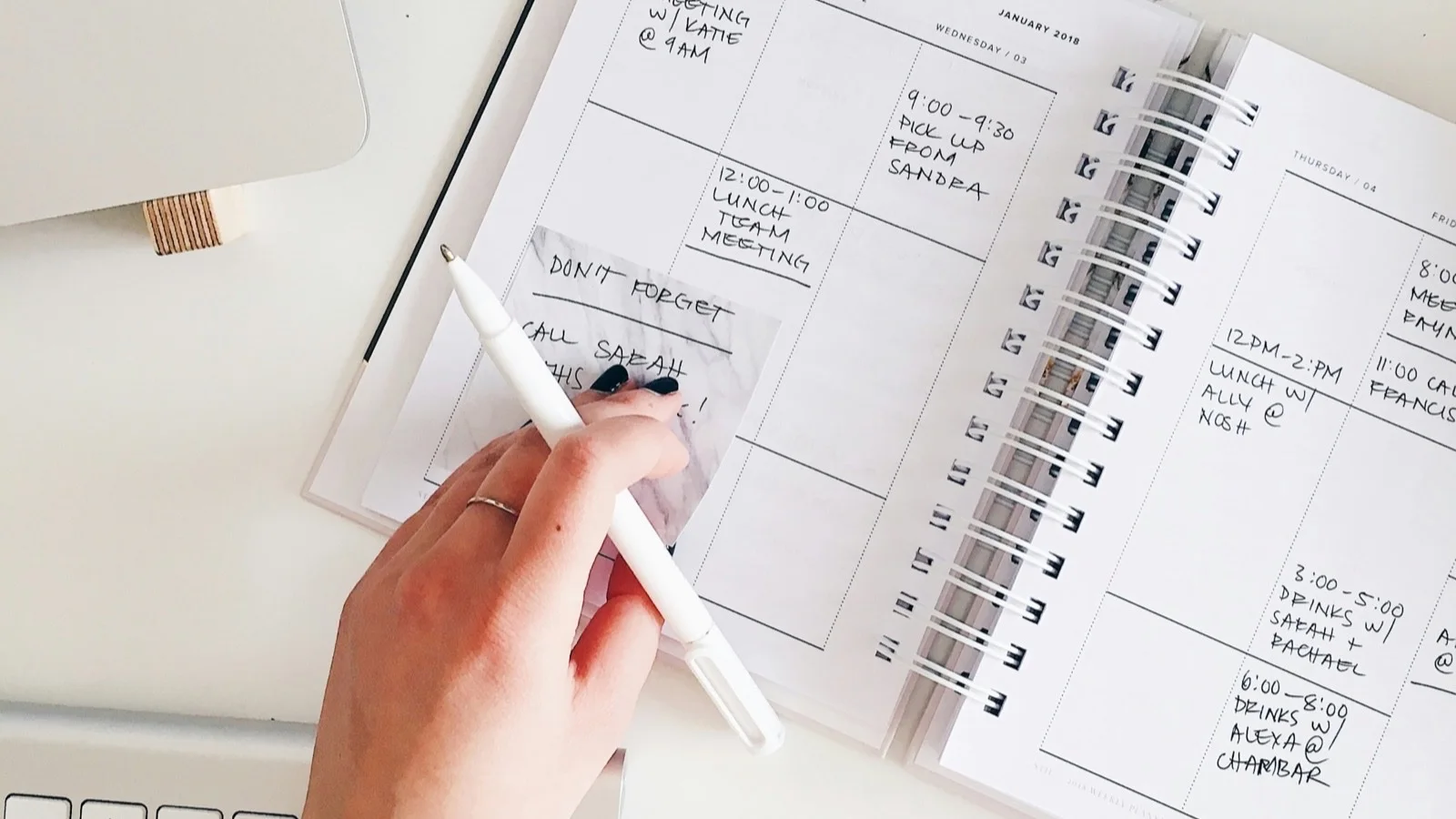 Main tenant un stylo au-dessus d'un carnet ouvert sur un bureau, avec ordinateur portable en arrière-plan.