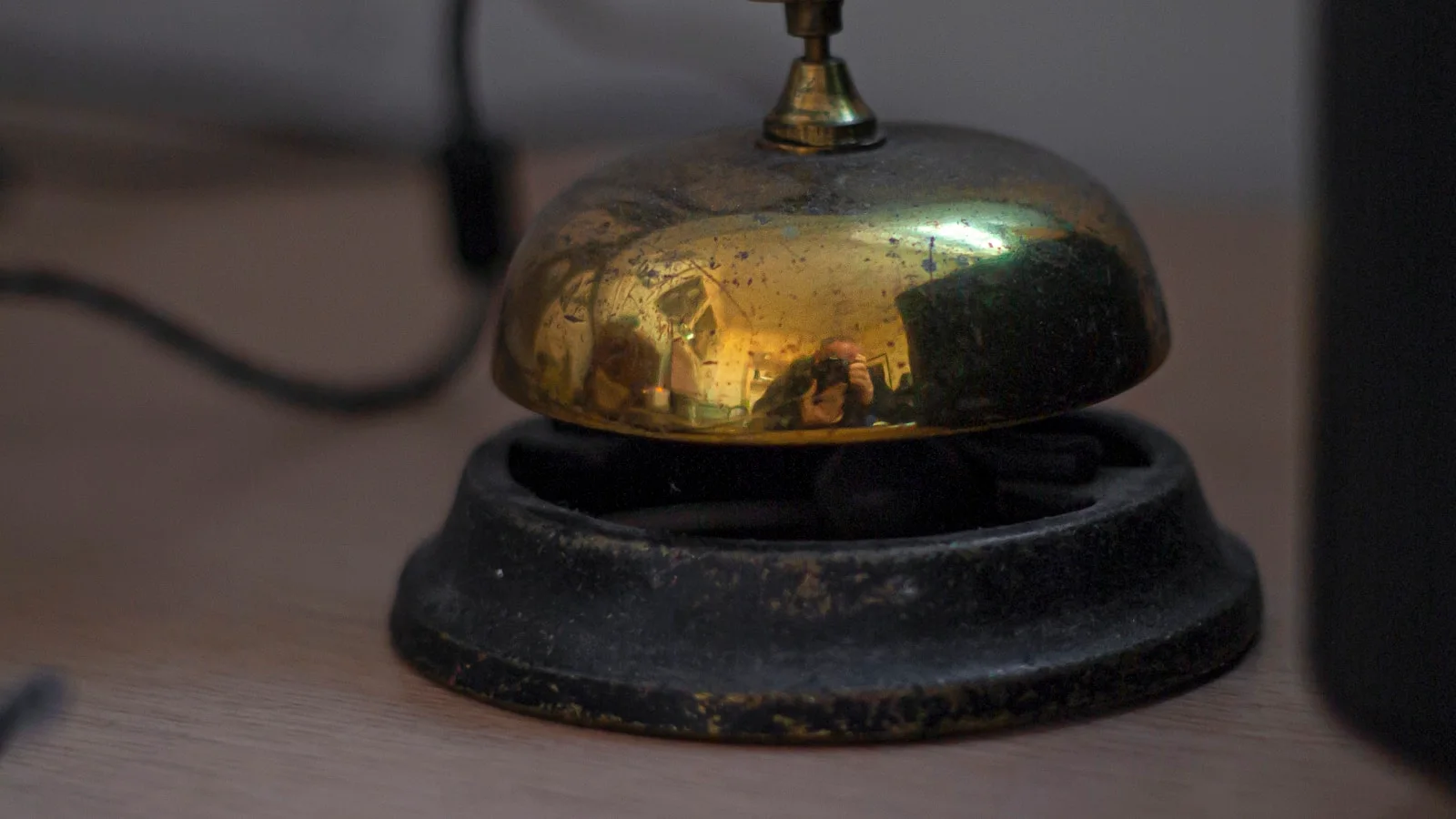 Sonnette de comptoir en laiton patiné posée sur un bureau en bois, l'objet qui appelle un humain au service, dans une lumière chaude.