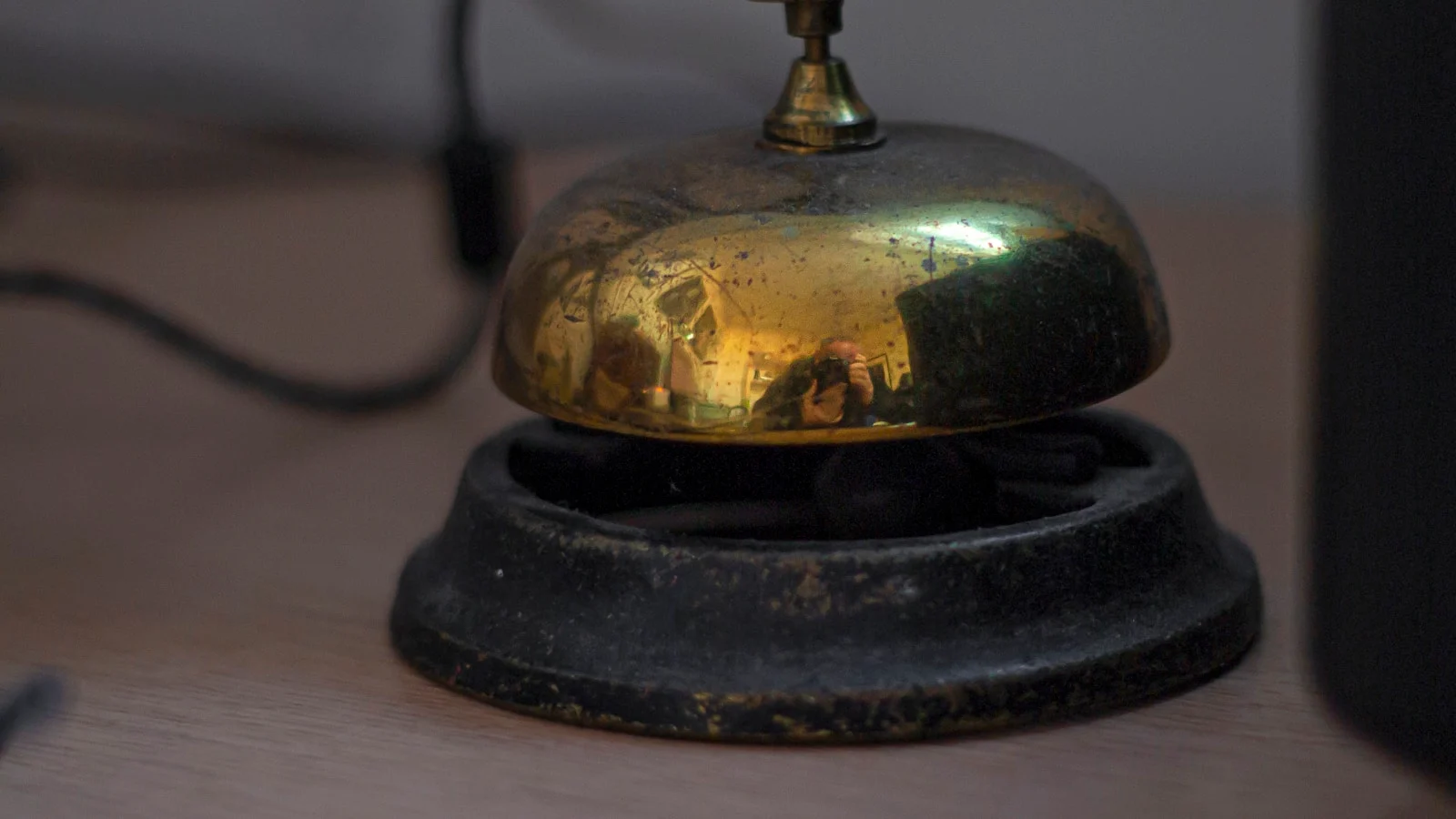 Sonnette de comptoir en laiton patiné posée sur un bureau en bois, l'objet qui appelle un humain au service, dans une lumière chaude.